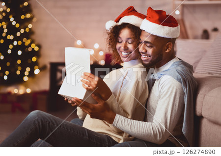 Sweet couple opening Christmas gift, wearing santa hats in front of xmas tree, free space Sweet couple opening Christmas gift, wearing santa hats in front of xmas tree, free space 126274890