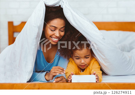 African American Mother And Daughter Using Mobile Phone Lying In Bed Covered With Blanket Indoor. Selective Focus 126274911