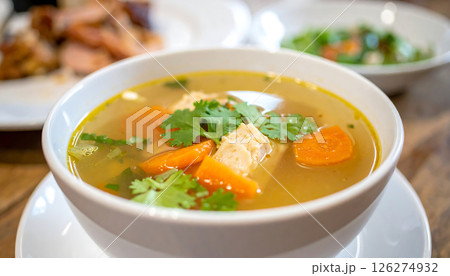 A bowl of chicken and carrot soup garnished with cilantro is served on a white plate. A bowl of chicken and carrot soup garnished with cilantro is served on a white plate. 126274932