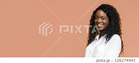 A happy black woman with curly, black hair is smiling while wearing a white shirt. She stands against a peach-colored background and looks directly at the camera. 126274942