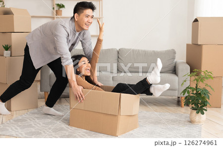 Home, People, Moving And Real Estate Concept. Excited asian couple having fun and riding in cardboard boxes in new flat 126274961