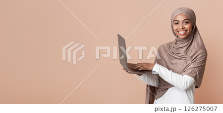Online education concept. Happy black muslim woman in hijab holding laptop, typing on keyboard and looking at camera, yellow background, free space Online education concept. Happy black muslim woman in hijab holding laptop, typing on keyboard and looking at camera, yellow background, free space 126275007