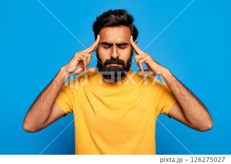 Indian man with a beard is shown with his eyes closed and his hands touching his temples, looking stressed and possibly in pain. He is wearing a yellow shirt against a blue background. 126275027