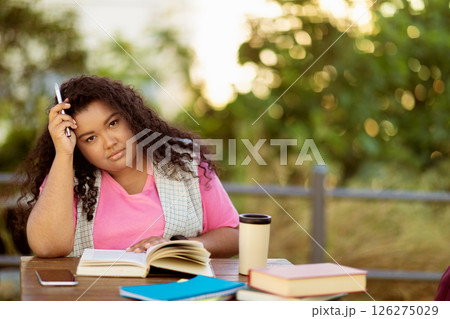 A student sits at a table surrounded by books, looking thoughtfully while holding a pen. She is outdoors in a sunny environment, deep in contemplation about her studies. 126275029