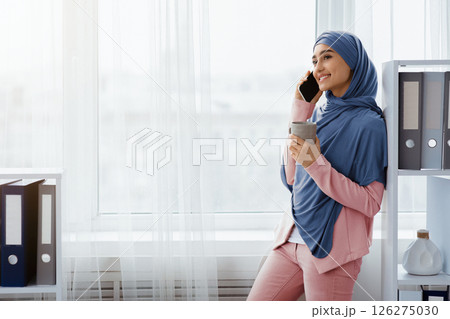 Young muslim businesswoman relaxing in office with smartphone and coffee, standing near window and holding mug with hot drink, copy space 126275030