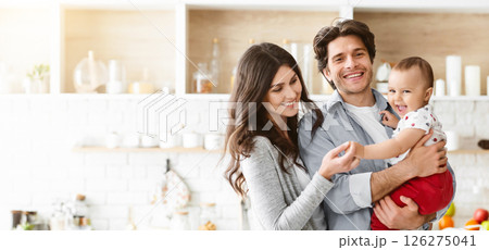 This image shows a happy family of three in a modern kitchen. The father is holding their baby, who is smiling and reaching out to the mother This image shows a happy family of three in a modern kitchen. The father is holding their baby, who is smiling and reaching out to the mother 126275041