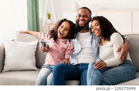 Relaxing Together. Portrait of happy young African American family watching television, parents sitting with their daughter on comfortable sofa in the living room, laughing at the program Relaxing Together. Portrait of happy young African American family watching television, parents sitting with their daughter on comfortable sofa in the living room, laughing at the program 126275049