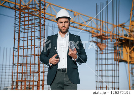 In hard hat, holding plan in hands. Businessman in formal clothes is on the construction site at daytime In hard hat, holding plan in hands. Businessman in formal clothes is on the construction site at daytime 126275192