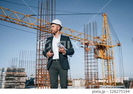 In hard hat, holding plan in hands. Businessman in formal clothes is on the construction site at daytime In hard hat, holding plan in hands. Businessman in formal clothes is on the construction site at daytime 126275194