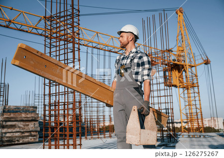 With big wooden plank in hand. Man is working on the construction site at daytime With big wooden plank in hand. Man is working on the construction site at daytime 126275267