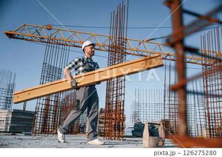 View from the side. Walking and holding wooden plank. Man is working on the construction site at daytime View from the side. Walking and holding wooden plank. Man is working on the construction site at daytime 126275280