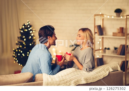 Young couple celebrating Christmas at home, relaxing with cups of coffee on sofa 126275302