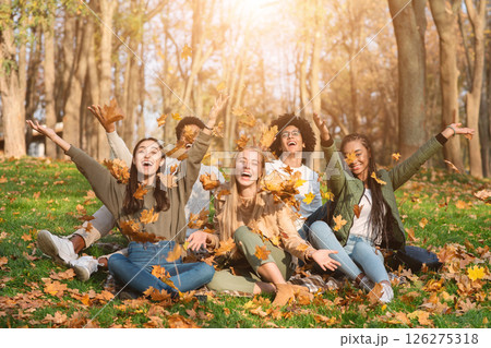 Group of cheerful international friends playing with autumn leaves in public park, taking autumn photos together 126275318