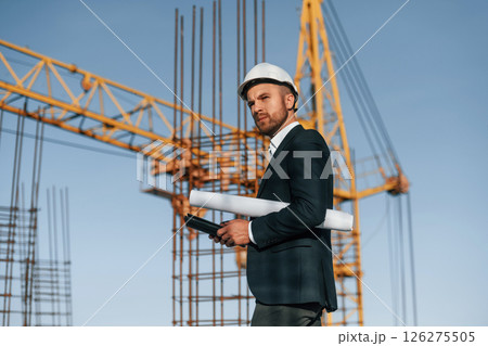 Side view. Walking and holding paper with plan and digital tablet. Businessman in formal clothes is on the construction site Side view. Walking and holding paper with plan and digital tablet. Businessman in formal clothes is on the construction site 126275505