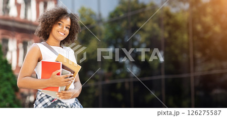 Student Orientation Concept. Portrait of happy black teen girl with backpack and workbooks standing outdoors in college campus, posing to camera 126275587