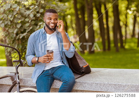 Handsome Afro Guy With Takeout Coffee Sitting On Bench In Park And Talking On Cellphone, Relaxing Outdoors 126275588