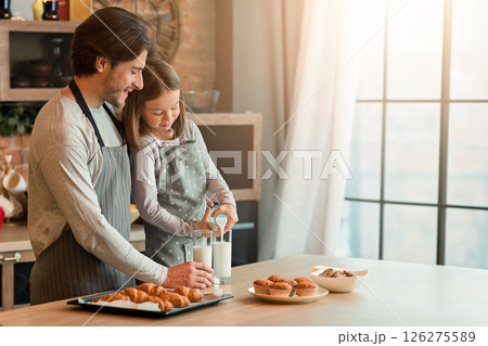 Little girl pouring milk to glasses while having bite with dad in kitchen, enjoying homemade pastry Little girl pouring milk to glasses while having bite with dad in kitchen, enjoying homemade pastry 126275589