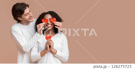 Romantic Surprise Concept. Loving guy covering eyes of his afro girlfriend with red paper heart cards, yellow studio background with empty space 126275725