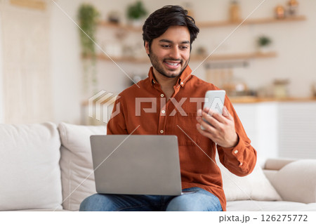 Arab man sits on a white couch in a living room. He is working on a laptop while looking at his phone. He is smiling, suggesting he is enjoying his work. Arab man sits on a white couch in a living room. He is working on a laptop while looking at his phone. He is smiling, suggesting he is enjoying his work. 126275772