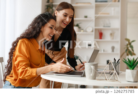 Two Happy Ladies Using Laptop Computer Working And Learning Online Together Or Making Video Call Sitting At Desk In Modern Office. E-Learning. Females Entrepreneurship Career Concept. Side View 126275785