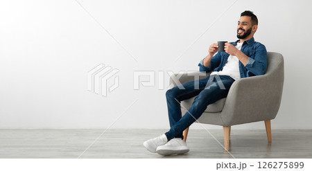 Full length of young Arab man drinking hot coffee in armchair against white studio wall, banner with free space. Peaceful middle Eastern guy having relaxing day, chilling on lazy morning Full length of young Arab man drinking hot coffee in armchair against white studio wall, banner with free space. Peaceful middle Eastern guy having relaxing day, chilling on lazy morning 126275899