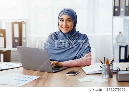 Modern Job For Muslim Women. Portrait of happy islamic girl in hijab working on laptop in office and looking at camera, copy space 126275962