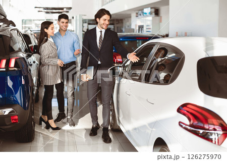 Car Rental Concept. Happy family choosing car for rent, manager showing them luxury auto standing in dealership store 126275970