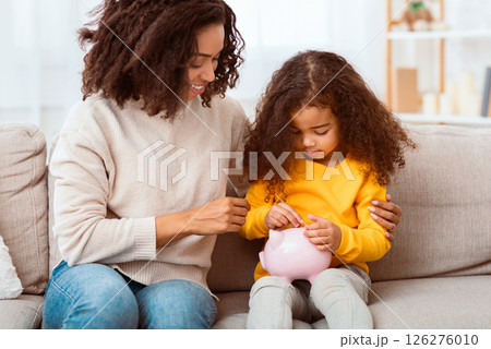 Personal Savings. Little African Girl Putting Money To Piggybank Sitting With Mother On Couch Indoor Personal Savings. Little African Girl Putting Money To Piggybank Sitting With Mother On Couch Indoor 126276010