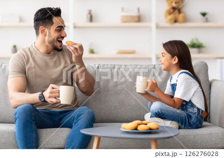 Snack Time. Portrait of cheerful little girl and man holding cups, drinking coffee and tea, eating cookies and looking at each other, dad and daughter talking sitting on the couch in living room 126276028