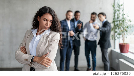 Workplace Sexism And Bullying. Unhappy Victimized Businesswoman Standing While Her Male Colleagues Whispering Behind Her Back Standing In Modern Office. Corporate Problems, Female Rights Concept 126276029