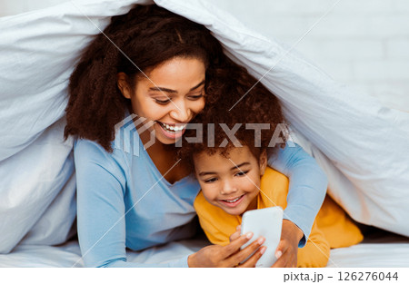 Happy Black Mother And Daughter Using Smartphone Lying In Bed Covered With Blanket In Bedroom. Selective Focus Happy Black Mother And Daughter Using Smartphone Lying In Bed Covered With Blanket In Bedroom. Selective Focus 126276044