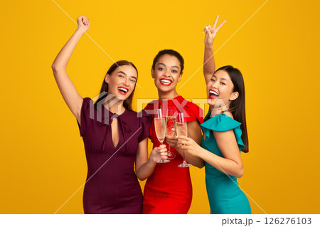 Glamour Party. Three Girls Models In Evening Dresses Clinking Glasses Having Fun Celebrating Something Standing Over Yellow Background 126276103