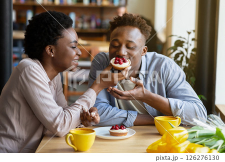 Lovely black woman feeding tasty berry tartlet to her boyfriend at cozy city cafe. Affectionate African American couple having dessert and coffee at restaurant, enjoying their first date Lovely black woman feeding tasty berry tartlet to her boyfriend at cozy city cafe. Affectionate African American couple having dessert and coffee at restaurant, enjoying their first date 126276130