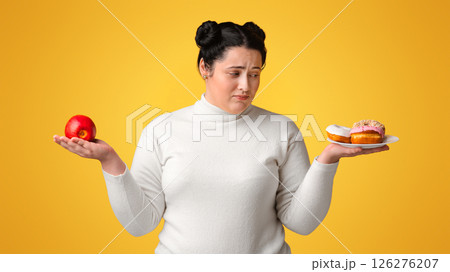Eat Or Not To Eat. Undecitive plump woman choosing between healthy and unhealthy food, holding apple and plate with donuts over yellow background 126276207