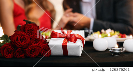 Romantic Couple Holding Hands, Celebrating Valentine's Day In Restaurant With Present Box And Red Roses Bouquet Lying On Table In Foreground, Closeup 126276261