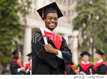 Cheerful african american guy in graduation costume showing his diploma and smiling at camera, black male student posing over international group of students at university campus, copy space Cheerful african american guy in graduation costume showing his diploma and smiling at camera, black male student posing over international group of students at university campus, copy space 126276332