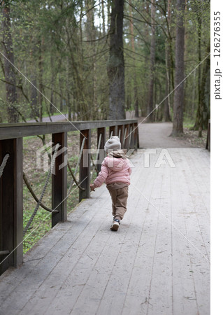 A small child happily runs along a wooden bridge in a quiet forest park, enjoying the beauty of spring, exploring nature and enjoying carefree moments of childhood. A small child runs along a bridge. 126276355