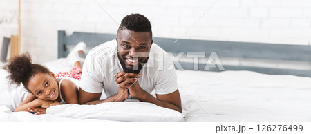African American father and his young daughter are lying playfully on a soft bed, smiling and enjoying their shared moments. The room has a bright and cheerful atmosphere, copy space African American father and his young daughter are lying playfully on a soft bed, smiling and enjoying their shared moments. The room has a bright and cheerful atmosphere, copy space 126276499