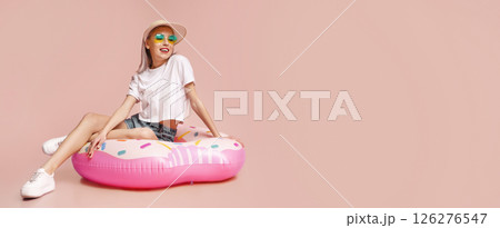 A young woman with pink hair is wearing sunglasses and a hat while sitting on a pink donut-shaped pool float. She is smiling and looking off to the side. The background is a solid pink color. 126276547