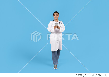 A healthcare professional Asian woman dressed in a white coat and scrubs walks confidently, engaged with a smartphone. The background is a vibrant blue, enhancing the professional's cheerful demeanor. A healthcare professional Asian woman dressed in a white coat and scrubs walks confidently, engaged with a smartphone. The background is a vibrant blue, enhancing the professional's cheerful demeanor. 126276564
