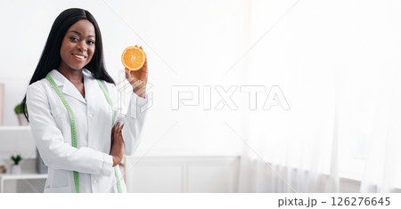 Start your day with fresh fruits. Positive black lady nutritionist holding orange half, standing in her office, copy space 126276645