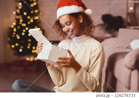 African-american girl looking inside Christmas present, sitting in front of xmas tree at home 126276733