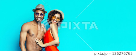 Smiling African American couple wearing sunglasses and hats in front of a bright blue background. The man is shirtless and the woman is wearing a red swimsuit, copy space 126276763