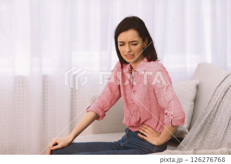 Side Stitch. Poor girl suffering from pain and cramps in her liver, sitting on sofa at home, holding spine, copyspace Side Stitch. Poor girl suffering from pain and cramps in her liver, sitting on sofa at home, holding spine, copyspace 126276768