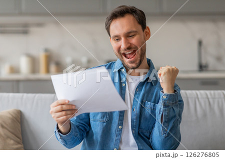 A young man sits on a couch in his home, reading a piece of paper with a big smile on his face. He is excited and holding his fist in the air. He is wearing a blue shirt over a white t-shirt. 126276805