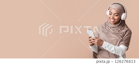 Cool music app. Smiling black muslim girl in hijab listening her favorite songs with wireless headphones online on smartphone Cool music app. Smiling black muslim girl in hijab listening her favorite songs with wireless headphones online on smartphone 126276813