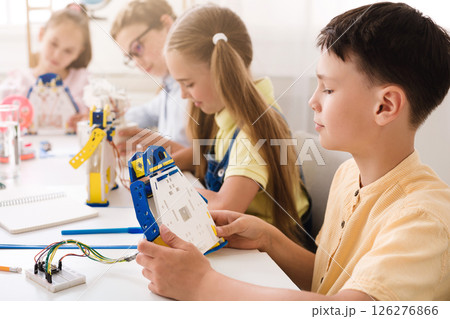 A young boy sits at a table in a classroom and builds a robot with other children. The boy is concentrating on his work as he holds a piece of the robot and examines it. 126276866