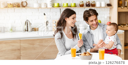A young family, consisting of a mother, father, and their baby, sit at a kitchen table enjoying breakfast. The mother is holding a piece of fruit in her hand and smiling at the camera, copy space 126276878