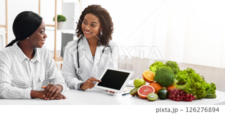 Friendly female dietologist showing black lady patient empty digital tablet screen in her office, technologies in medical care Friendly female dietologist showing black lady patient empty digital tablet screen in her office, technologies in medical care 126276894