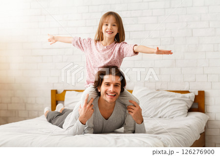 Father and daughter spending time together at home in morning. Happy little girl sitting on father shoulders with hands up, dad laying on bed, free space Father and daughter spending time together at home in morning. Happy little girl sitting on father shoulders with hands up, dad laying on bed, free space 126276900